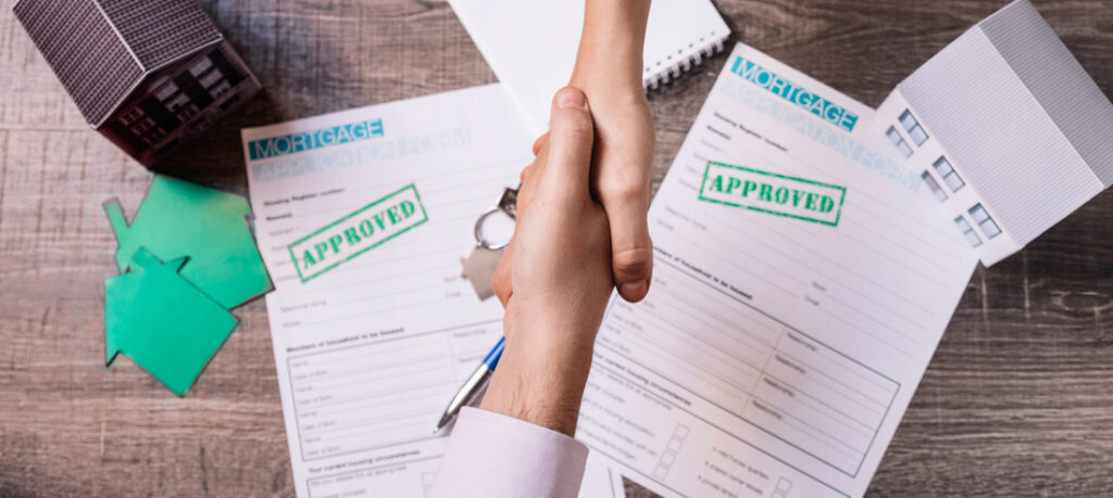 Handshake over approved mortgage application forms with house models and keys, symbolizing successful loan approval and real estate agreement.