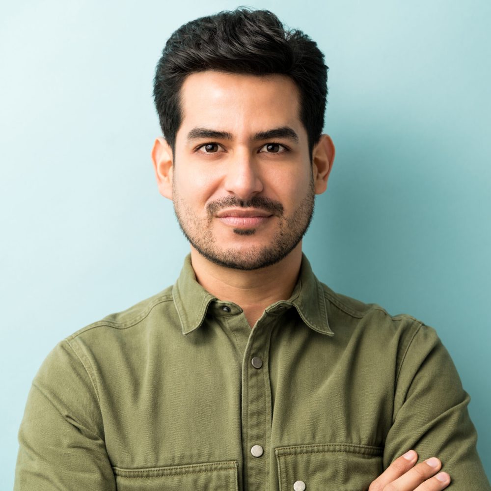Confident young man with neatly styled hair and a trimmed beard, standing against a light blue background with arms crossed.