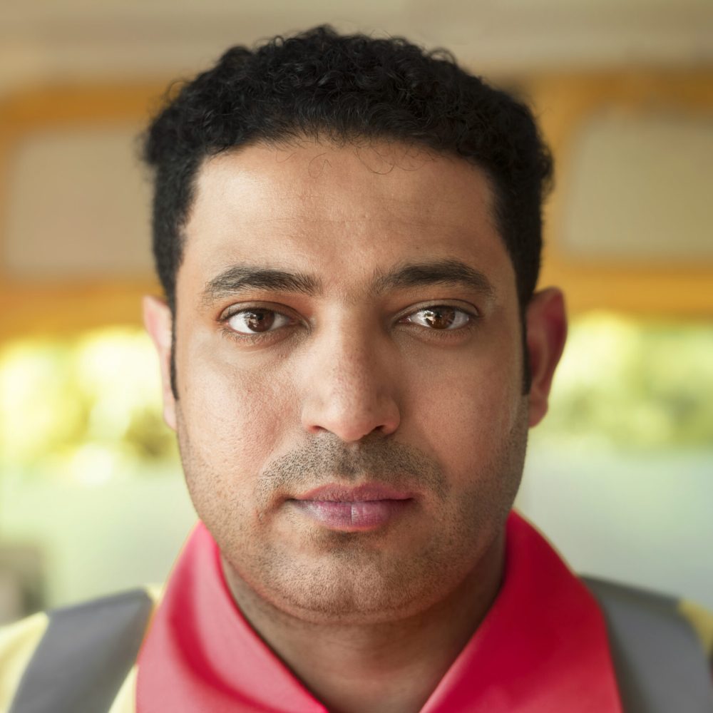 Man with short curly hair and serious expression, wearing a red collared shirt and backpack straps, with a soft blurred background.