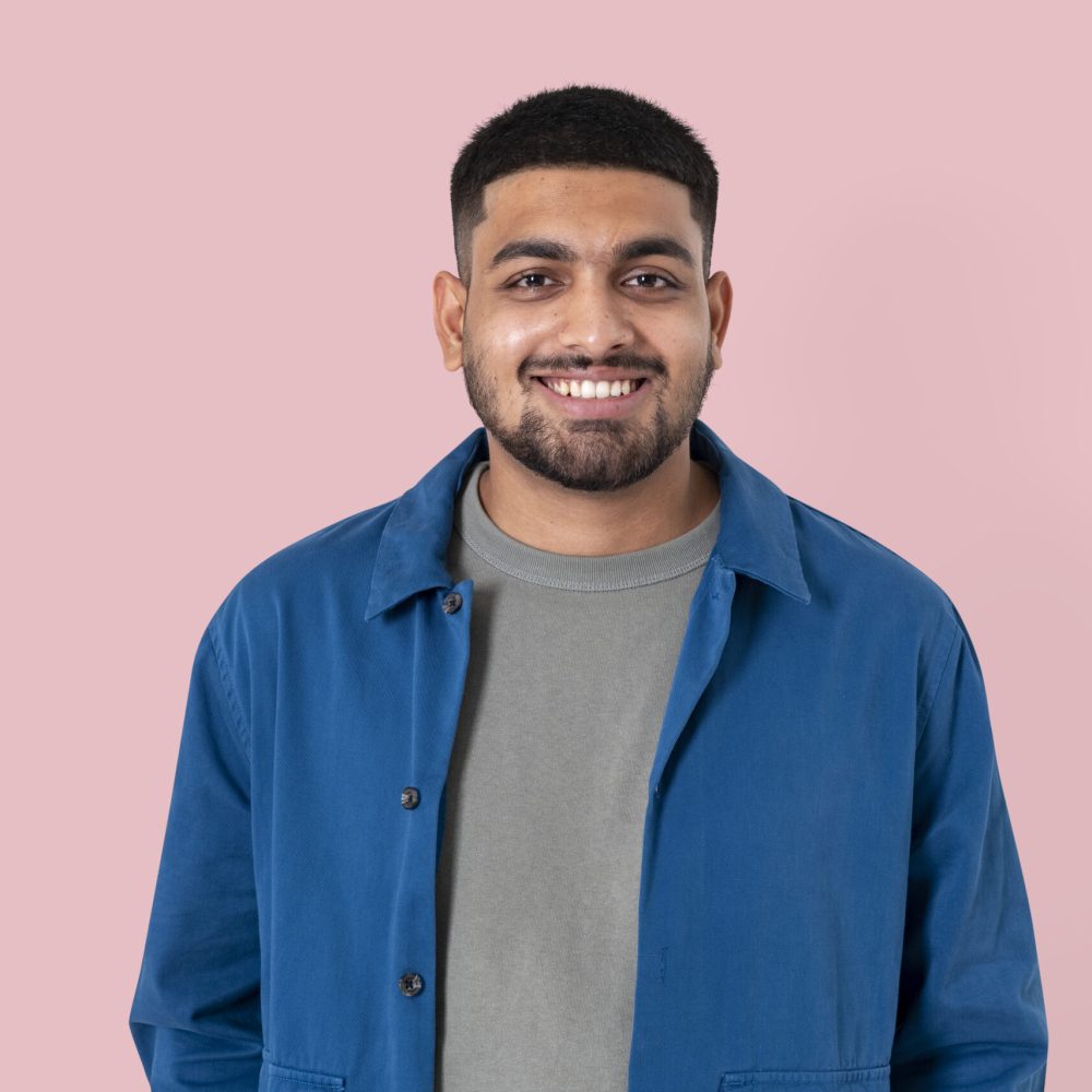Smiling young man in a blue jacket and grey t-shirt standing confidently against a soft pink background.