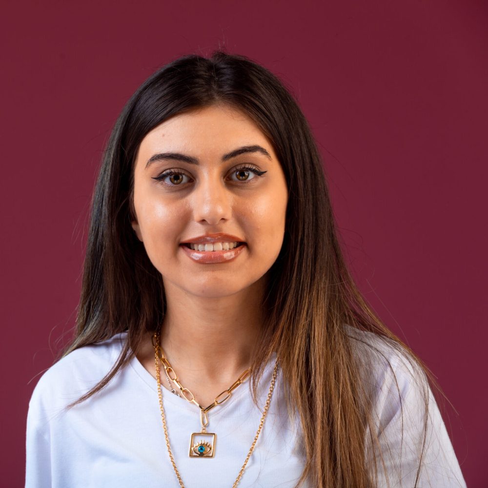 Smiling young woman with long straight hair, wearing a white top and gold necklaces, standing against a dark maroon background.
